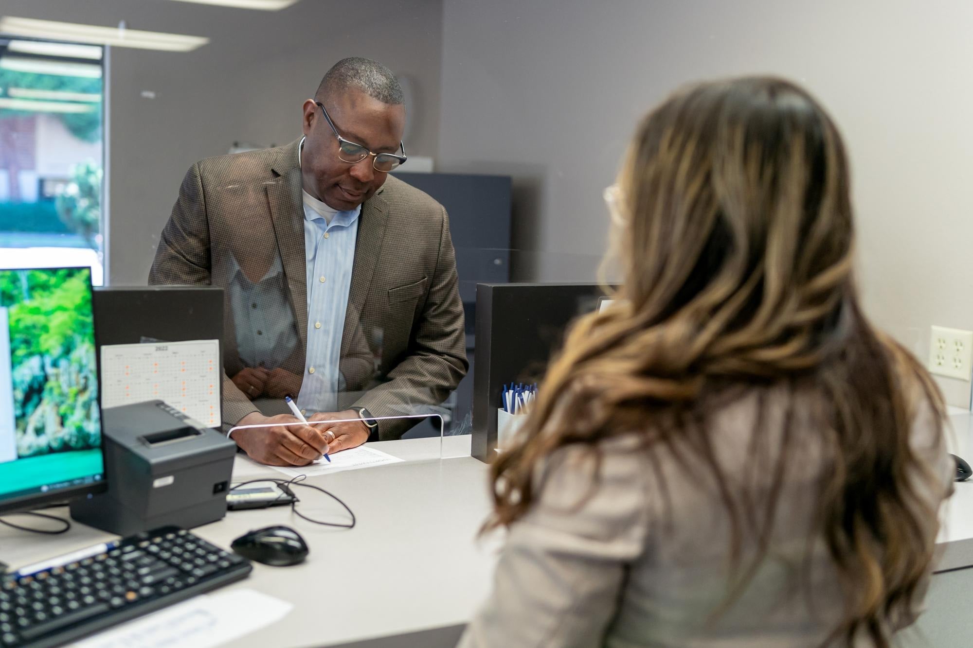 A visitor signing a paper register at a reception counter while the receptionist looks on